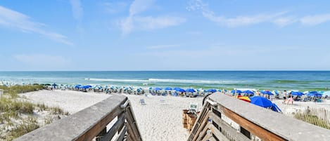 A wooden boardwalk leading to a beach with rows of blue umbrellas and beach chairs.
