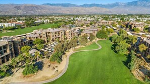 Vista aérea de un complejo turístico con varias edificaciones, un campo de golf y terreno montañoso al fondo.