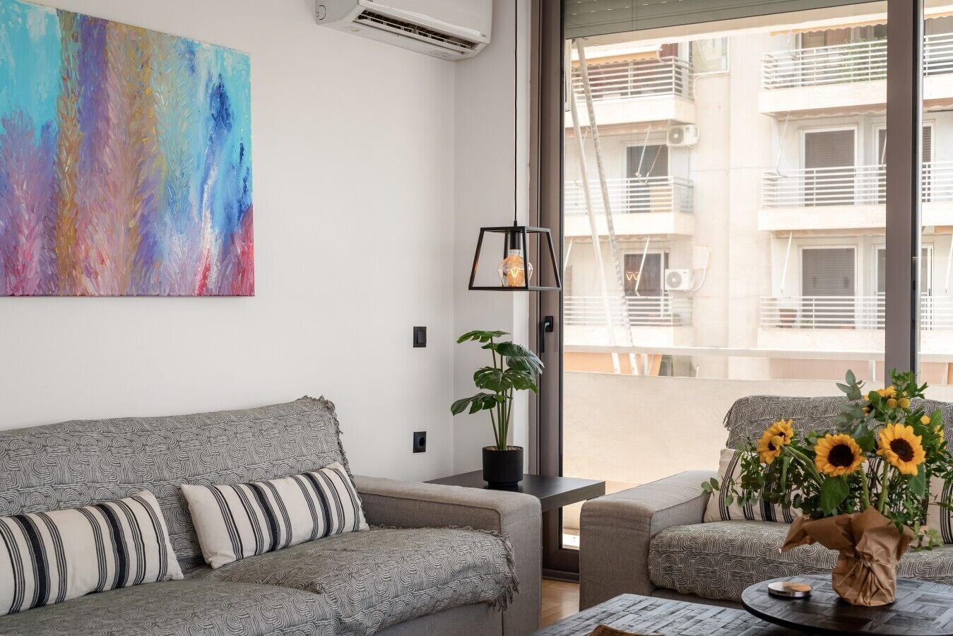 A modern living room with a grey sofa, a colorful abstract painting, a potted plant, and a view of a building through a large window.