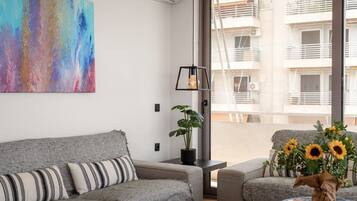 A modern living room with a grey sofa, a colorful abstract painting, a potted plant, and a view of a building through a large window.