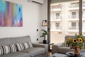 A modern living room with a grey sofa, a colorful abstract painting, a potted plant, and a view of a building through a large window. - Seafront 3BR with Panoramic Balcony & Parking (Paleo Faliro)