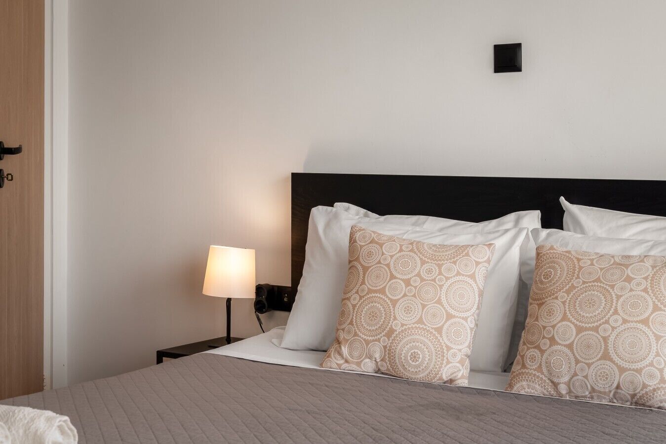 A hotel room with a bed, a nightstand with a lamp, and a wall-mounted control panel.