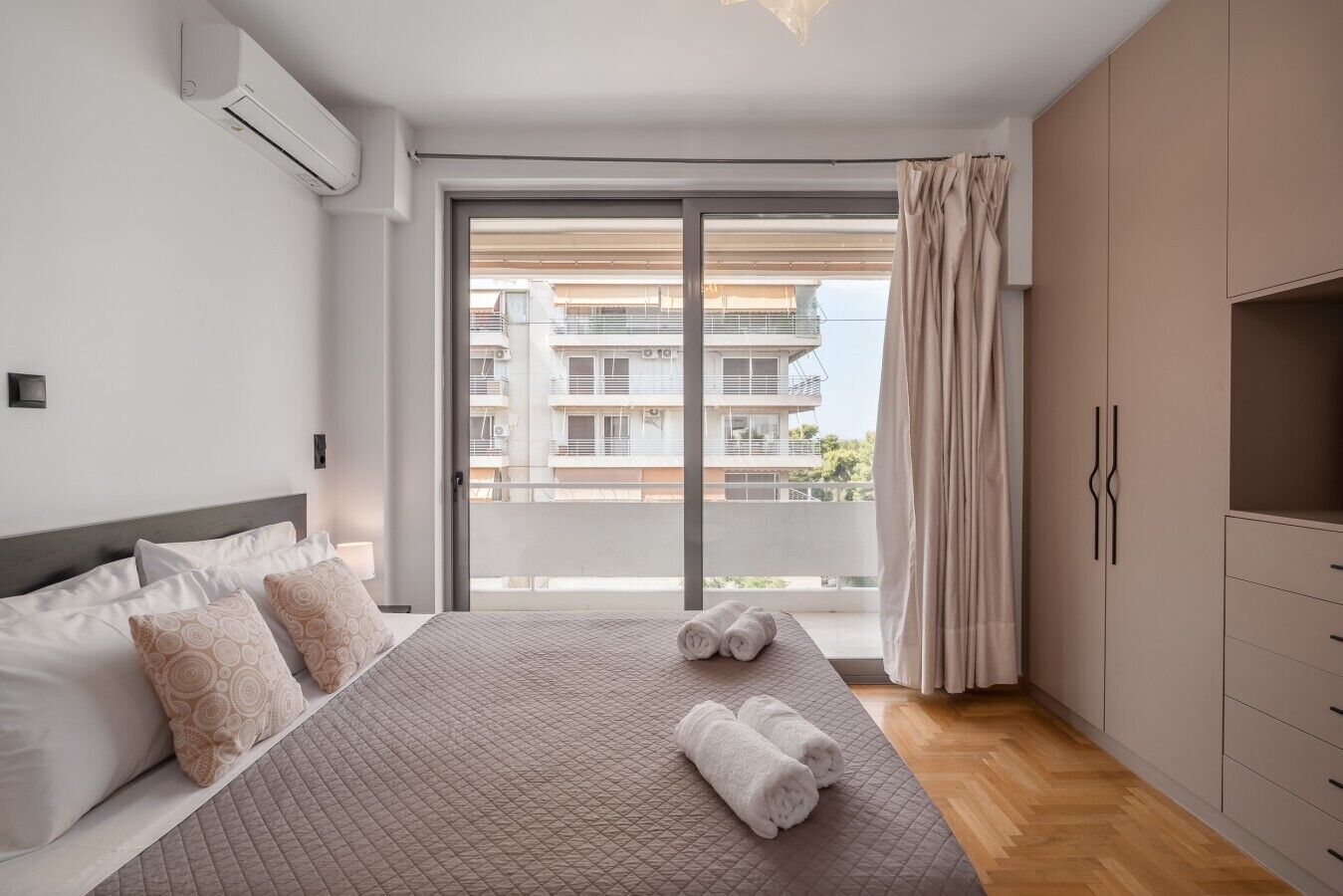 A hotel room with a bed, a wardrobe, a window with curtains, and a view of buildings outside.