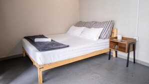 Een enkel bed met houten frame, witte beddengoed en gestreepte kussens. Er staat een nachtkastje met een lamp en een klein plankje.