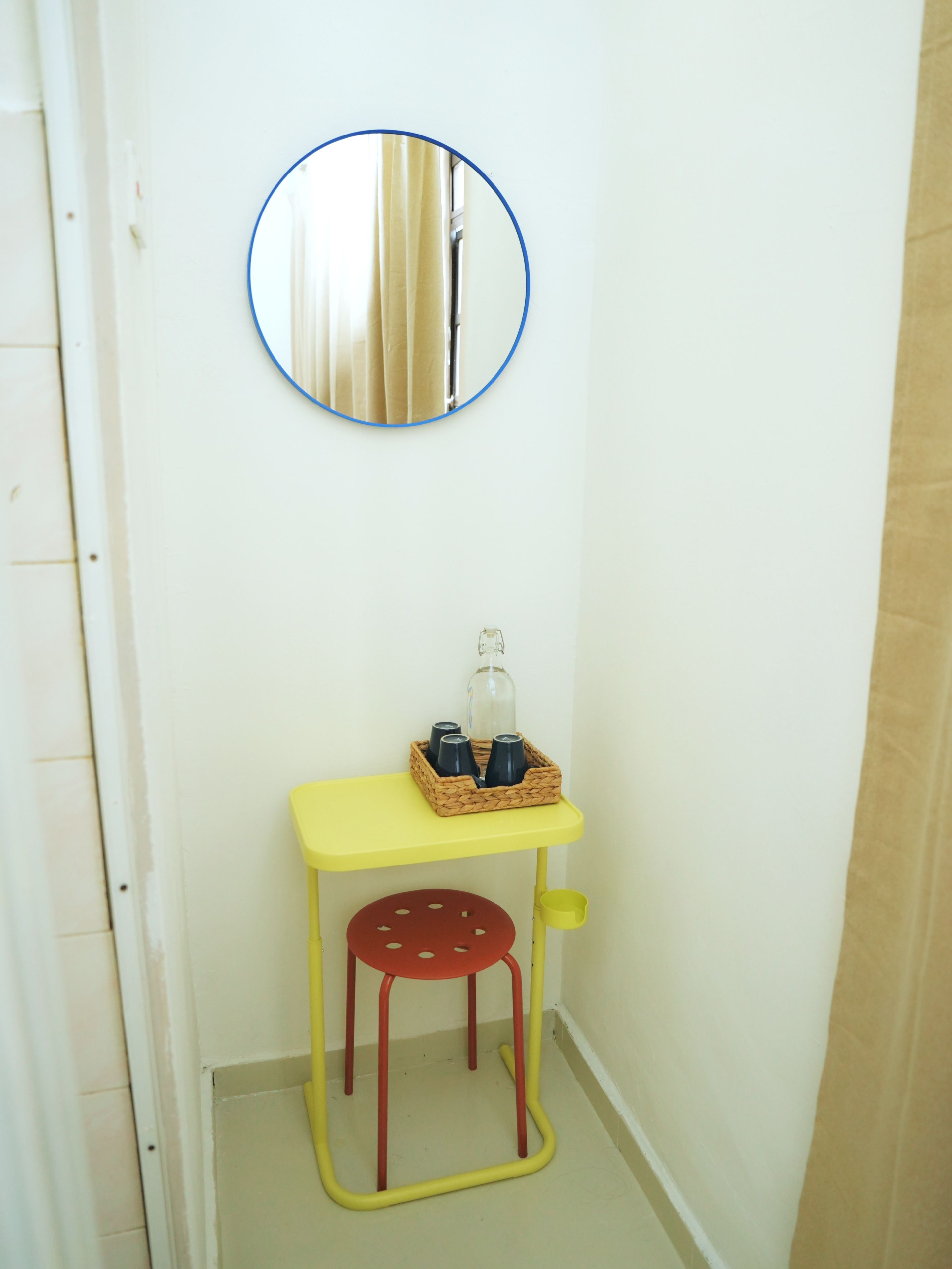 Un baño pequeño con un taburete amarillo, un espejo y una cesta de mimbre con tazas.