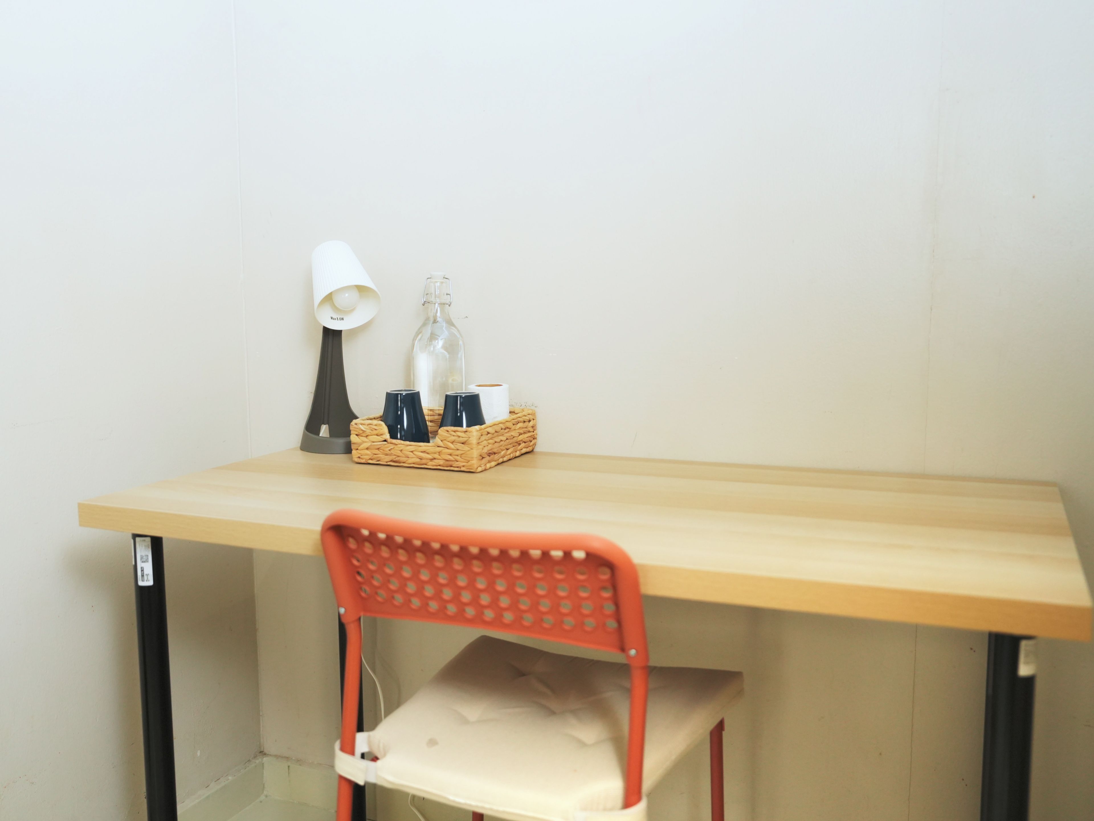 Un pequeño espacio de trabajo con un escritorio de madera, una silla roja, una lámpara y una cesta de mimbre con tazas y una botella.