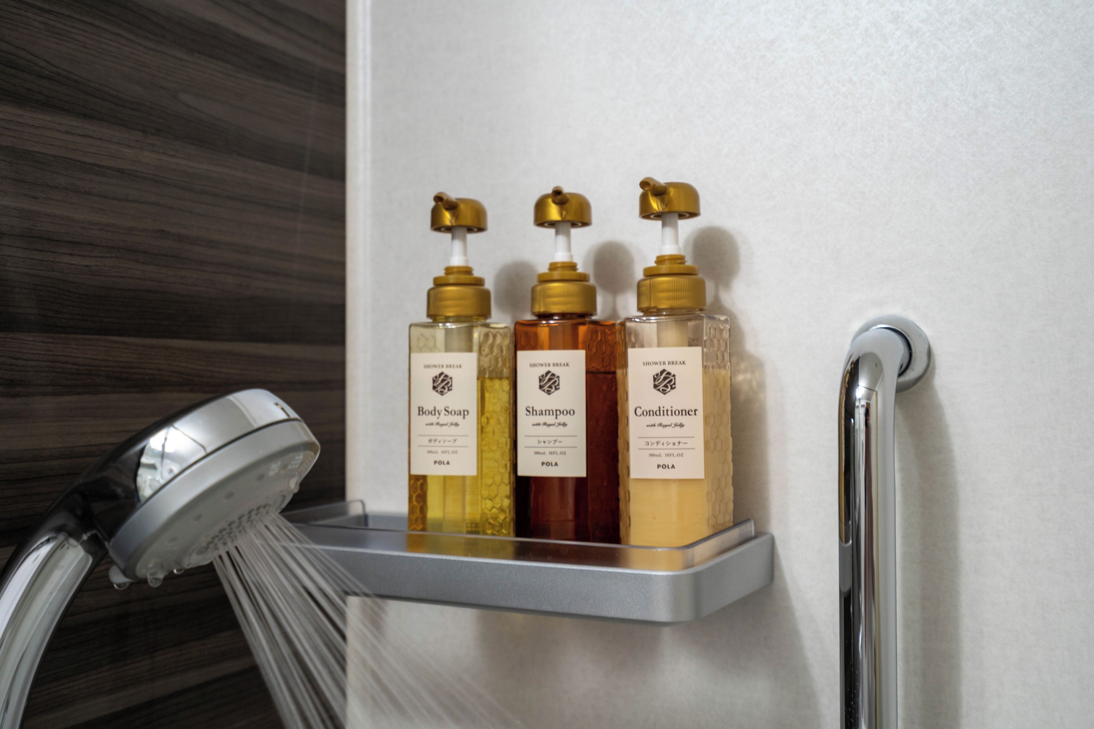 Une douche avec trois flacons de savon pour le corps, de shampoing et d’après-shampoing, placés sur une étagère fixée au mur.