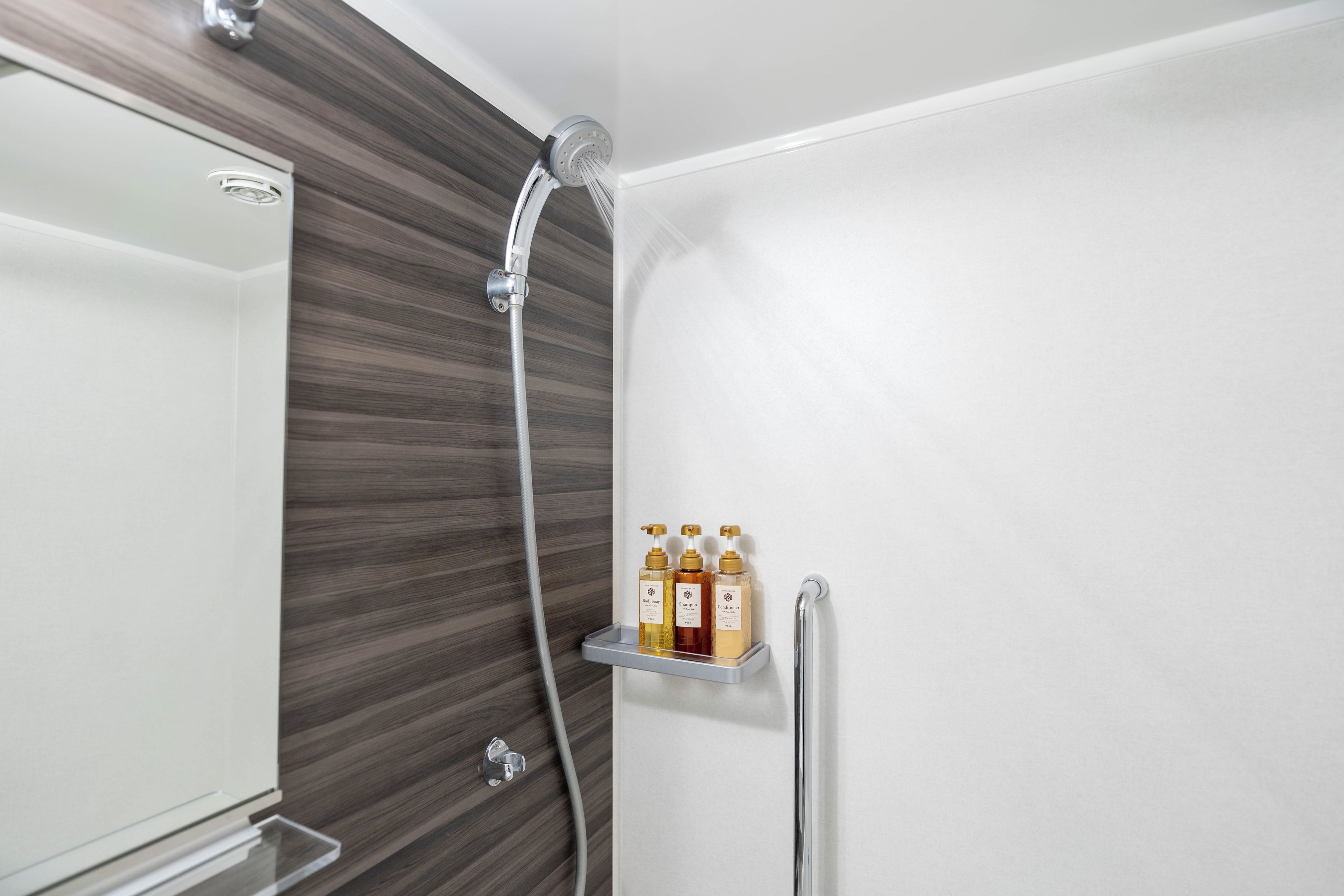 A shower with a handheld showerhead and a wall-mounted faucet. There is a shelf with three bottles of toiletries.