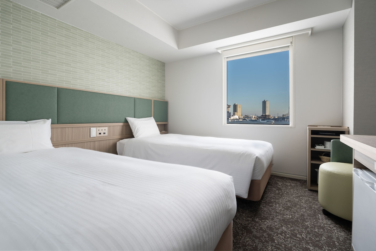 A hotel room with two beds, a window with a city view, a desk, and a chair.
