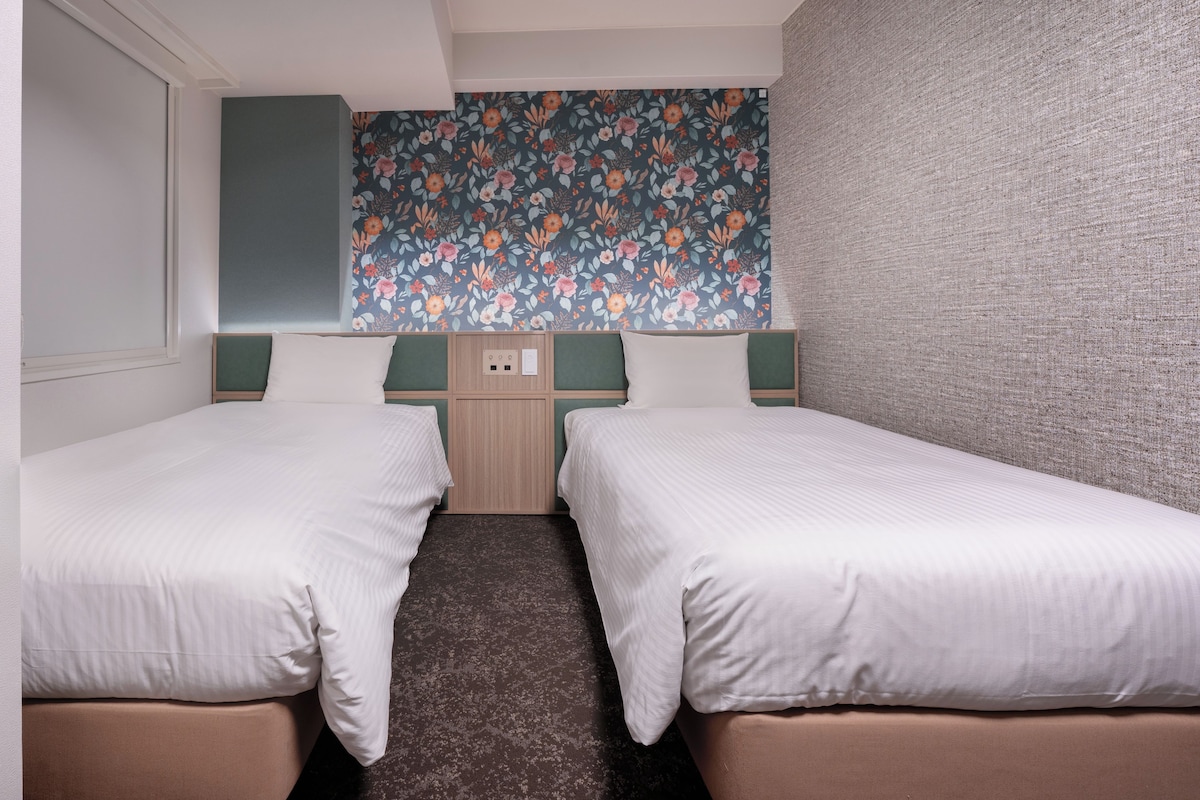 A hotel room with two single beds, a floral-patterned wall, and a textured grey wall.