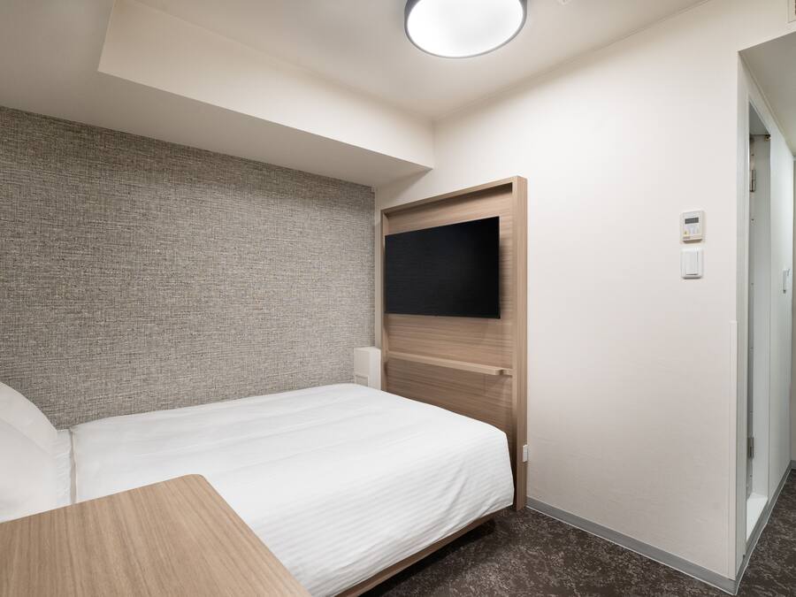 A hotel room with a bed, a flat-screen TV mounted on the wall, a door, and a light switch.