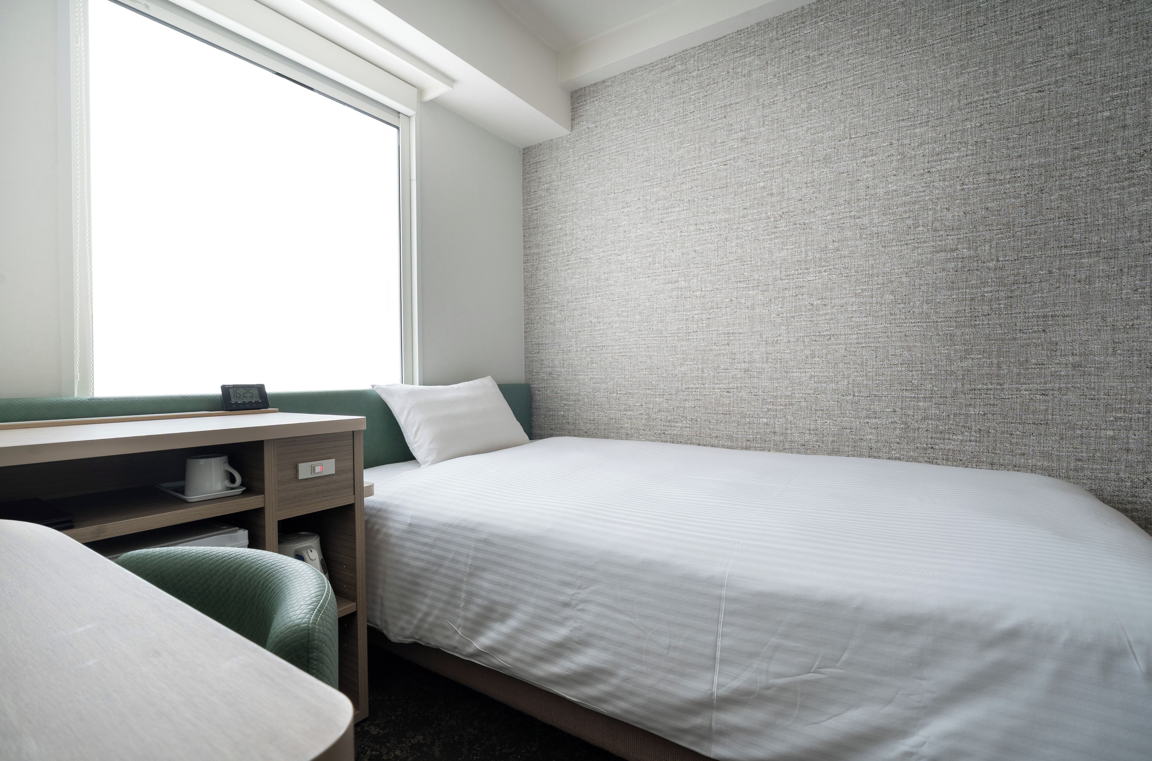 Quarto de hotel com uma cama de solteiro, uma escrivaninha com cadeira, uma mesinha e uma janela.