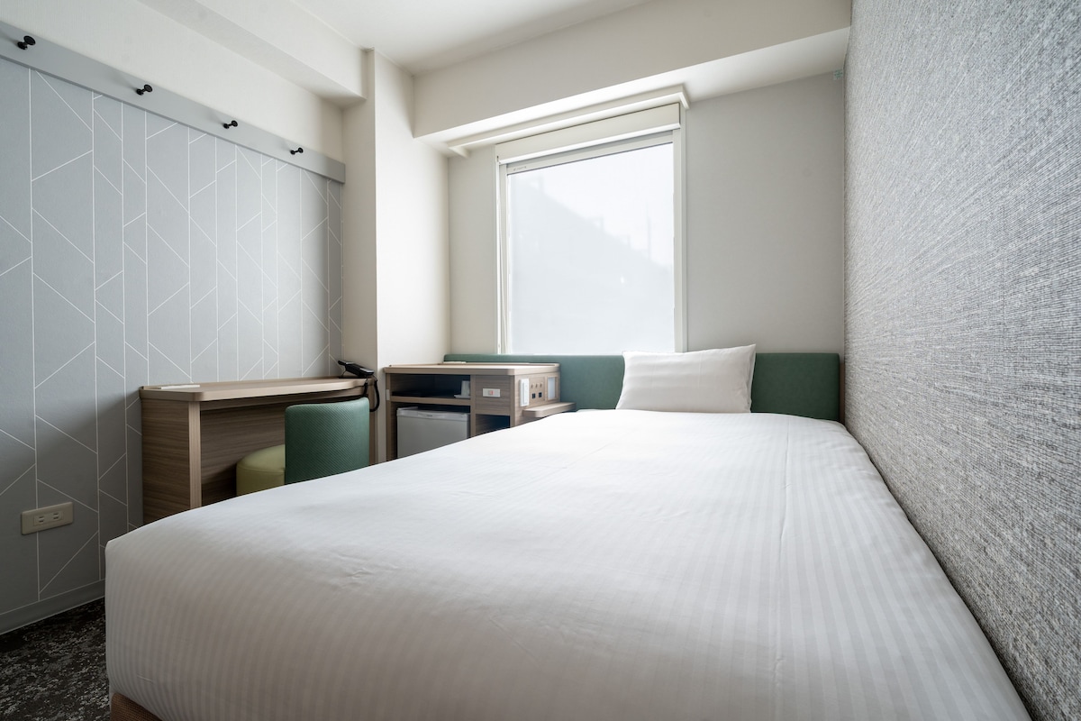 A hotel room with a bed, a desk, a chair, a window, and a wardrobe.