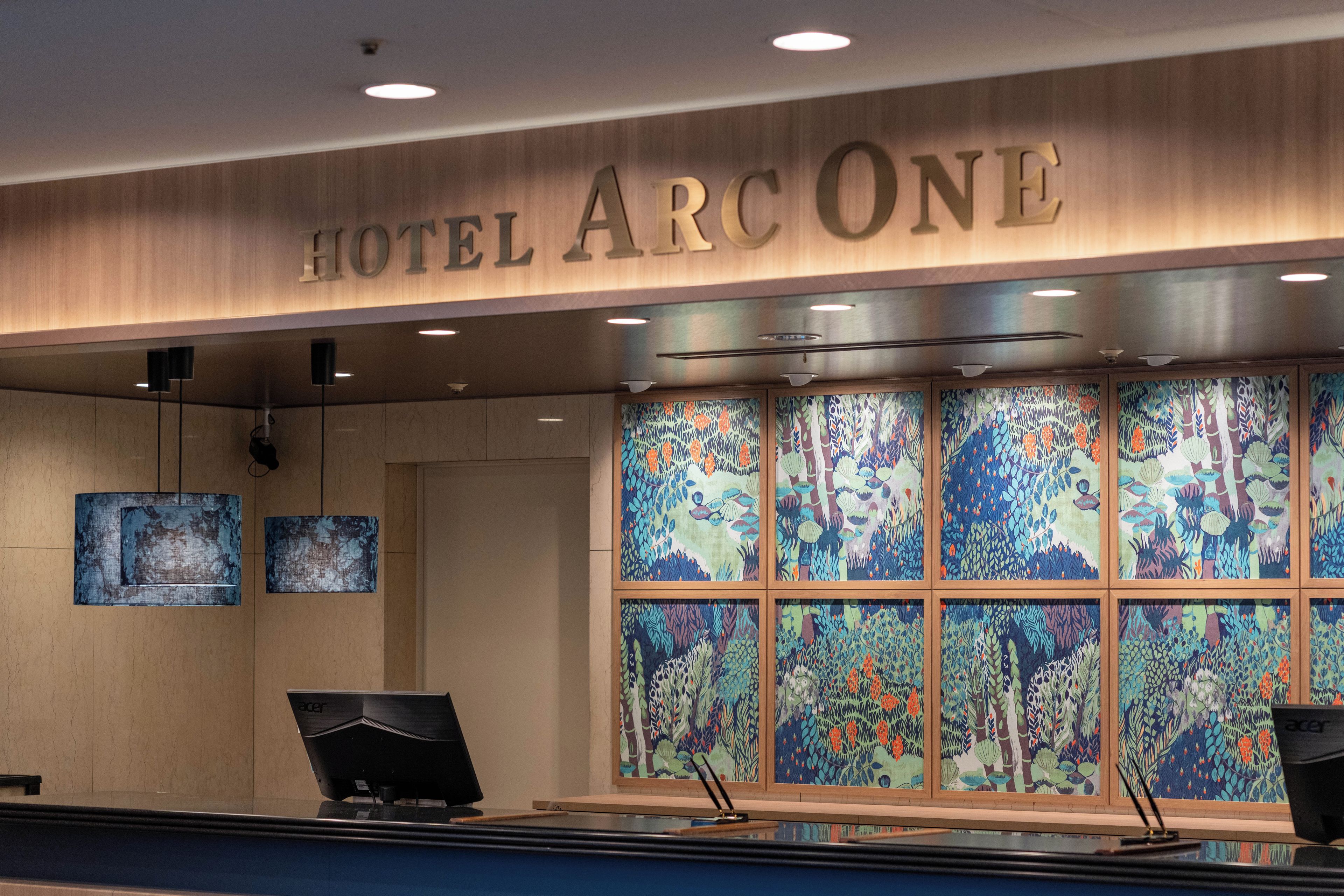 Un mostrador de recepción de hotel con un cartel que dice 'HOTEL ARC ONE'. Hay dos monitores sobre el mostrador y una ventana grande con arte colorido detrás.