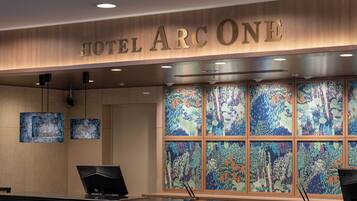 Un mostrador de recepción de hotel con un cartel que dice 'HOTEL ARC ONE'. Hay dos monitores sobre el mostrador y una ventana grande con arte colorido detrás.