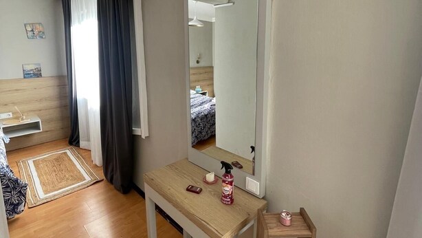 A hotel room with a wooden floor, a small table with a mirror, a bedside table, a shelf, and a window with curtains. - Cozy 1-bedroom flat in near Old Town with AC and balcony (Antalya)