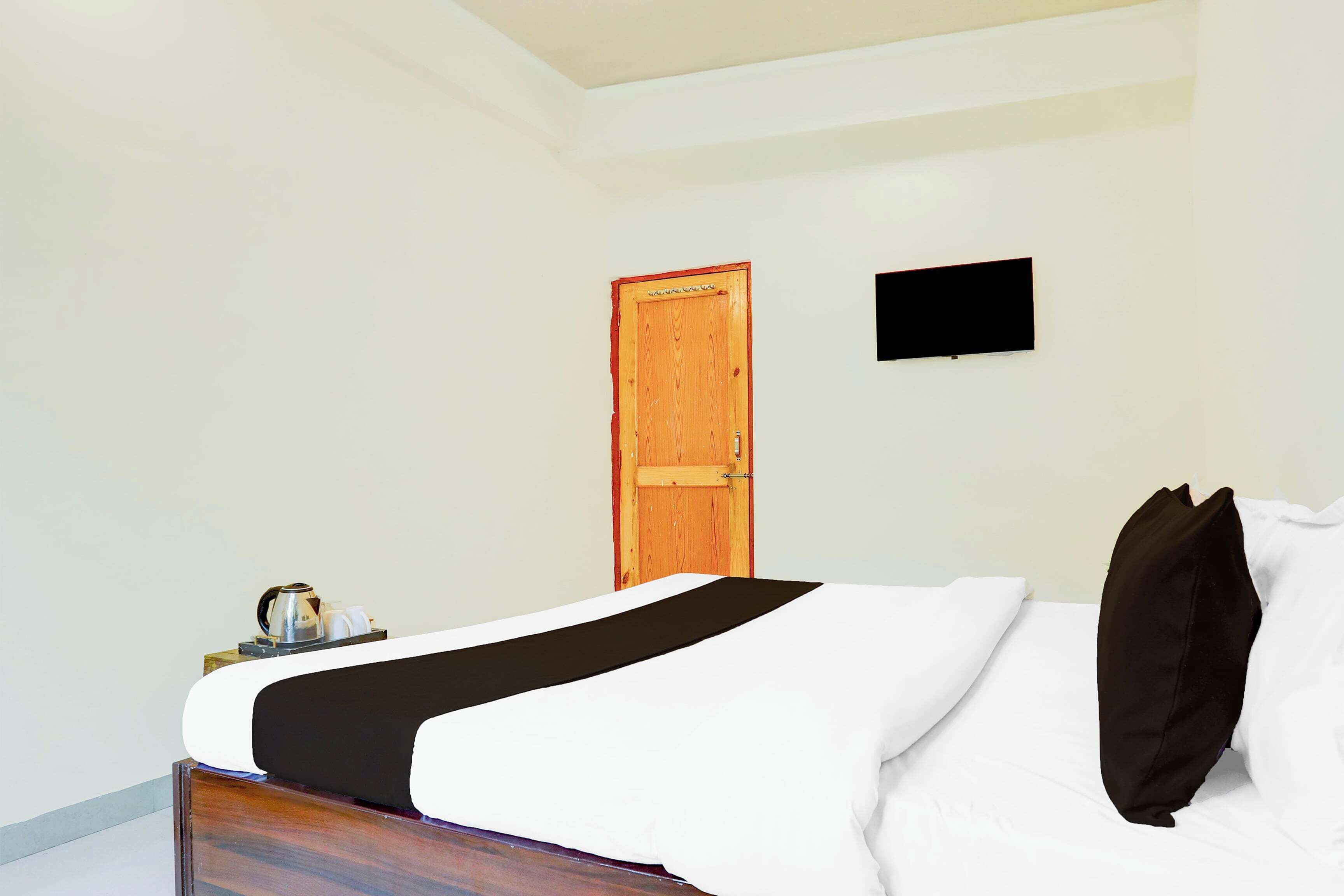 A hotel room with a bed, a wooden door, a small table with a kettle, and a flat-screen TV mounted on the wall.