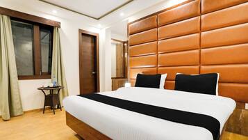 Una habitación de hotel con una cama grande, cabecera de madera, una mesita con una botella y una ventana con cortinas.