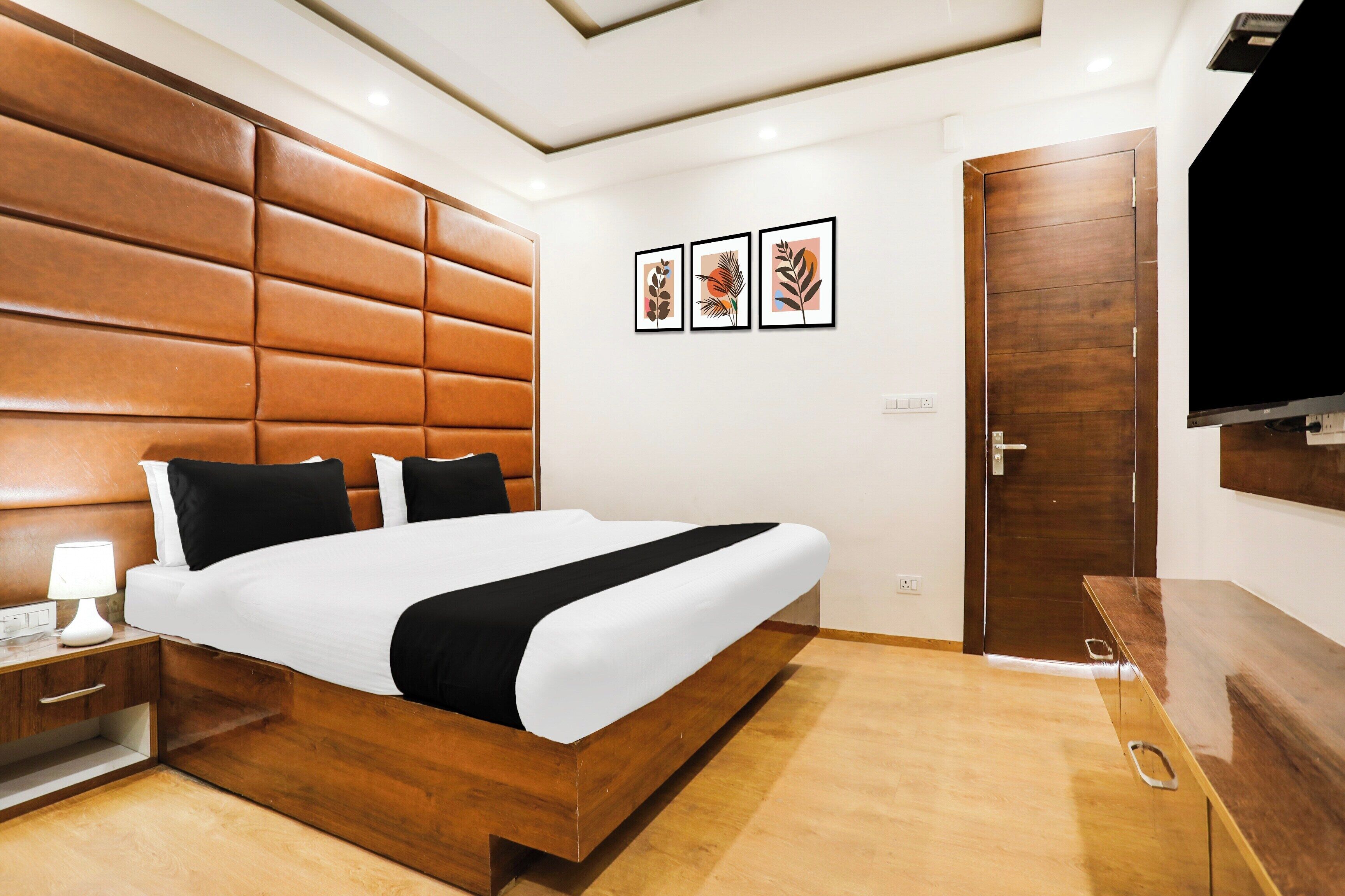 A modern hotel room with a wooden headboard, a large bed, a bedside table with a lamp, and a flat-screen TV mounted on the wall.