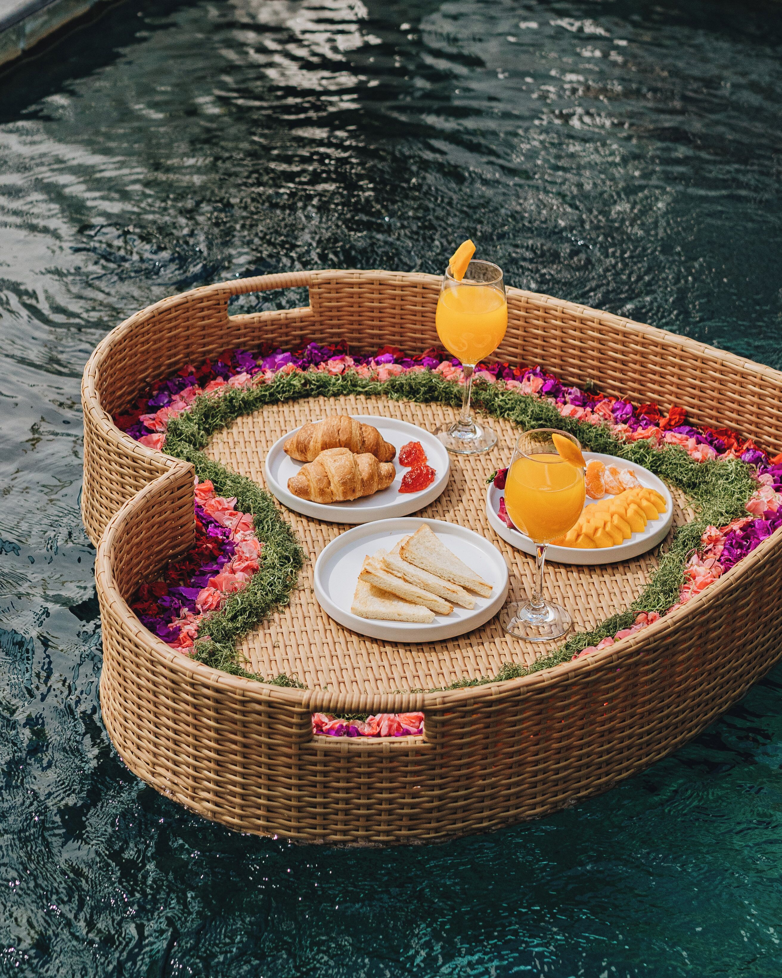 A wicker boat with plates of food and drinks, floating on water.