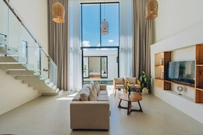 Ein modernes Wohnzimmer mit einem großen Sofa, einem Couchtisch und einem Fernseher auf einem Holzregal. Eine Treppe mit Glasgeländern führt zu einem Balkon.