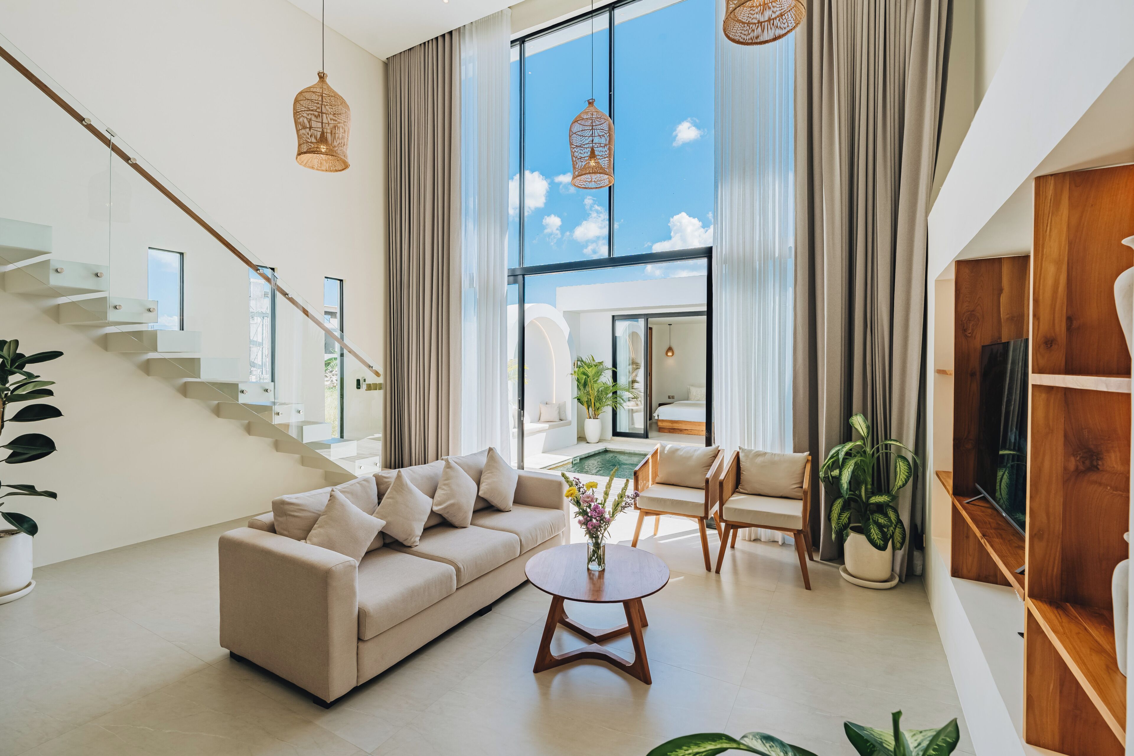 A spacious living area with a modern staircase, a beige sofa, a round coffee table, and large windows offering a view of the sky.