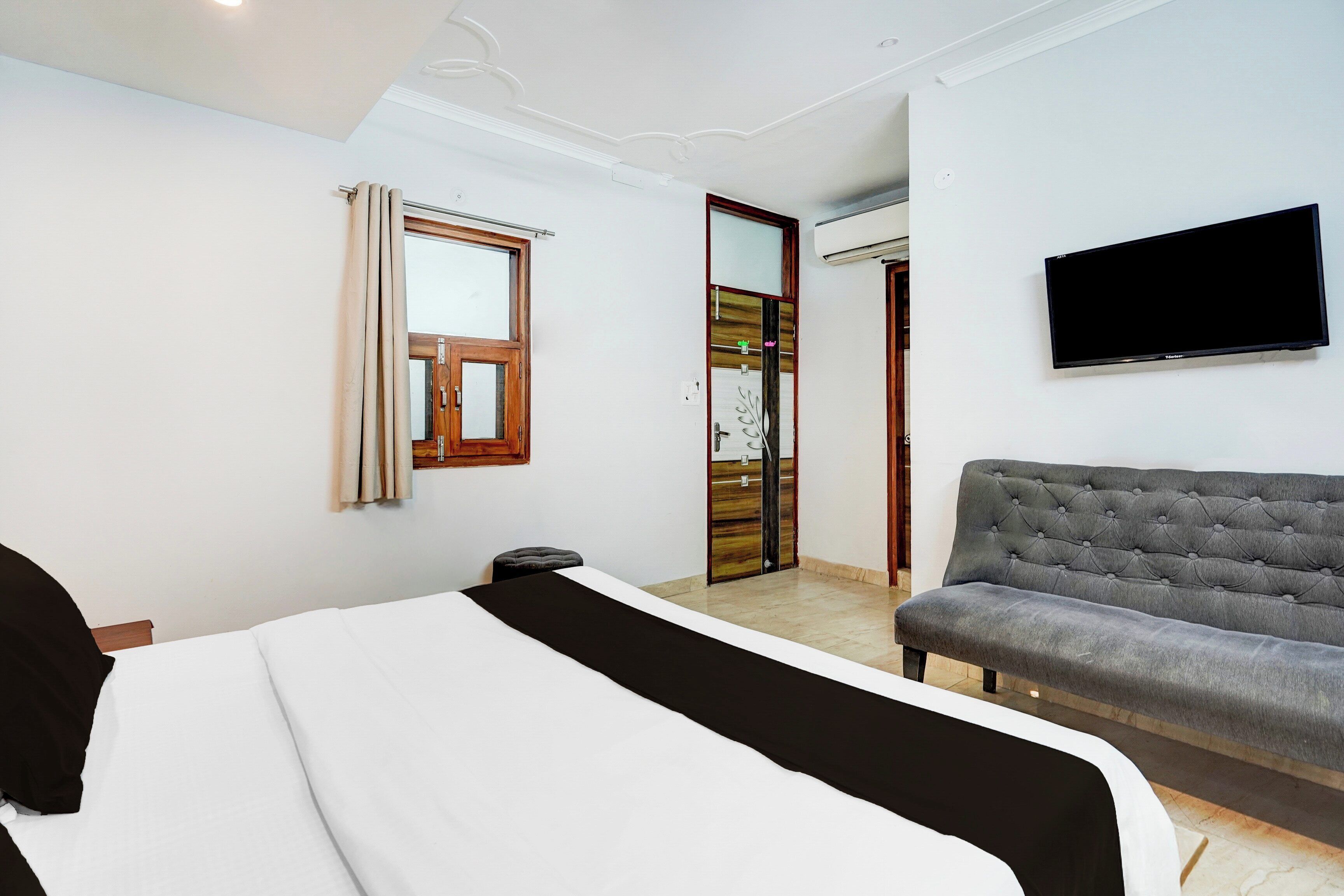 Una habitación de hotel con cama, sofá gris, televisión de pantalla plana, ventana con cortinas y puerta con panel decorativo.