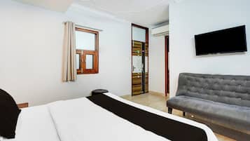 Una habitación de hotel con cama, sofá gris, televisión de pantalla plana, ventana con cortinas y puerta con panel decorativo.