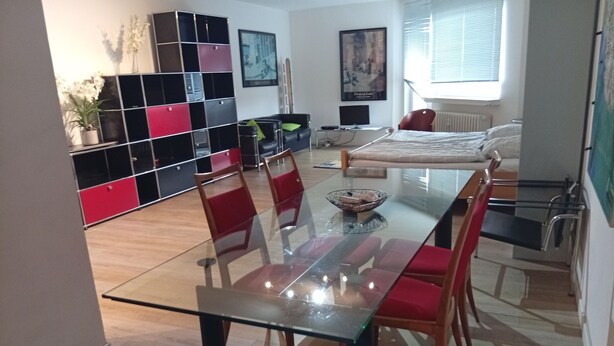 Ein modernes Wohnzimmer mit einem Glastisch, roten Stühlen, einer Couch und einem schwarzen, roten Regalelement.