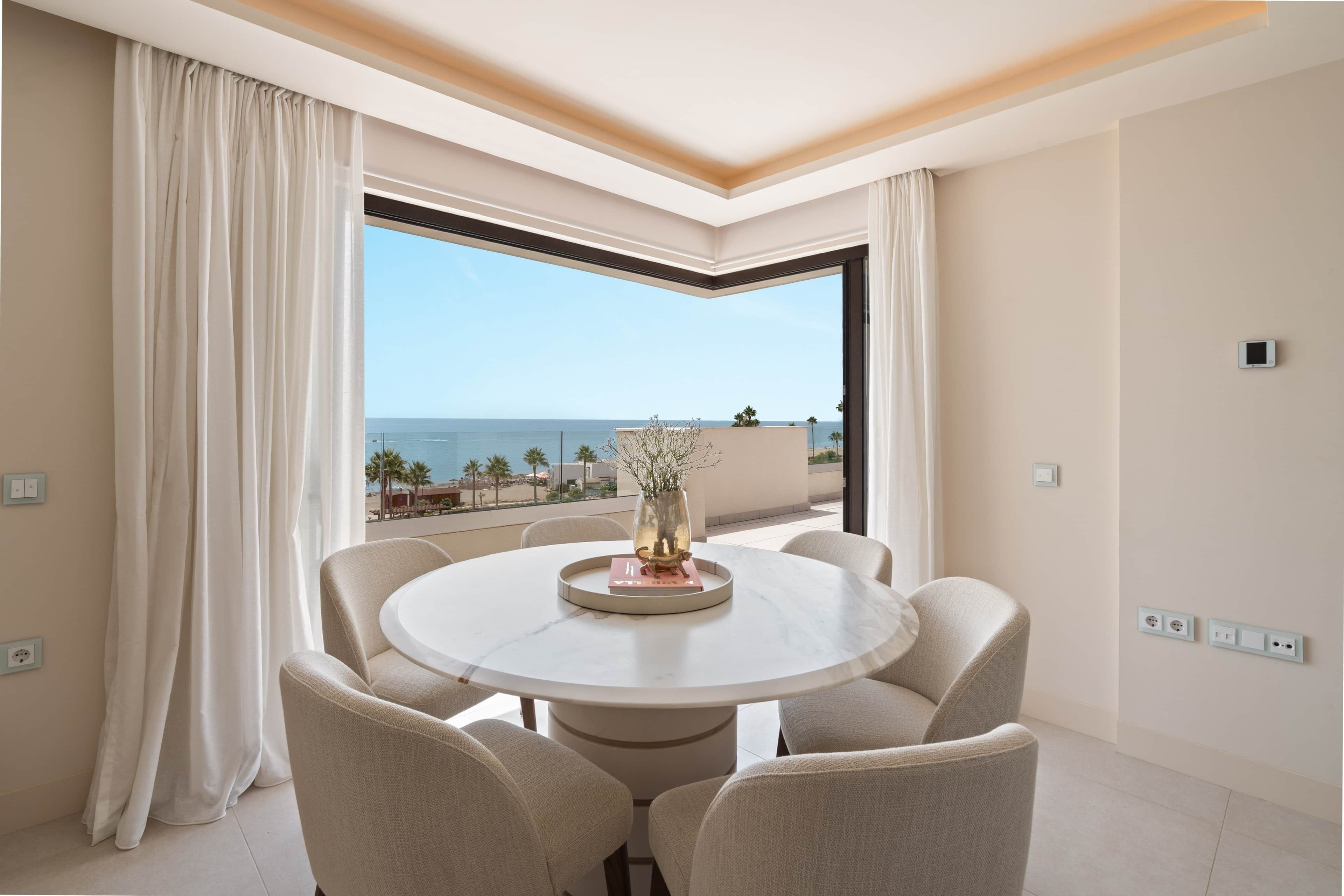 A round dining table with six chairs, a vase with a plant, and a view of the sea through a large window.