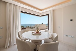 Ein runder Esstisch mit sechs Stühlen, eine Vase mit Pflanze und Blick auf das Meer durch ein großes Fenster.