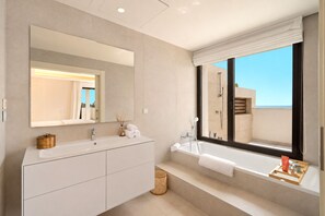 Une salle de bain moderne avec un grand miroir, deux lavabos, une baignoire et une fenêtre donnant sur la mer.