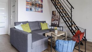 Ein modernes Wohnzimmer mit einem grauen Sofa, einem Couchtisch und einem schwarzen Metallstuhl mit blauer Sitzkissen. Im Hintergrund ist eine Treppe mit schwarzen Geländern zu sehen.