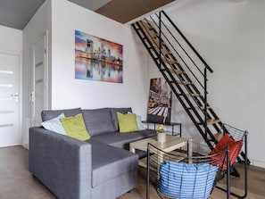A modern living room with a grey sofa, a coffee table, and a black metal chair with a blue cushion. A staircase with black railings is visible in the background. - House in Saint-pabu Near Stunning Beaches (Warsaw)