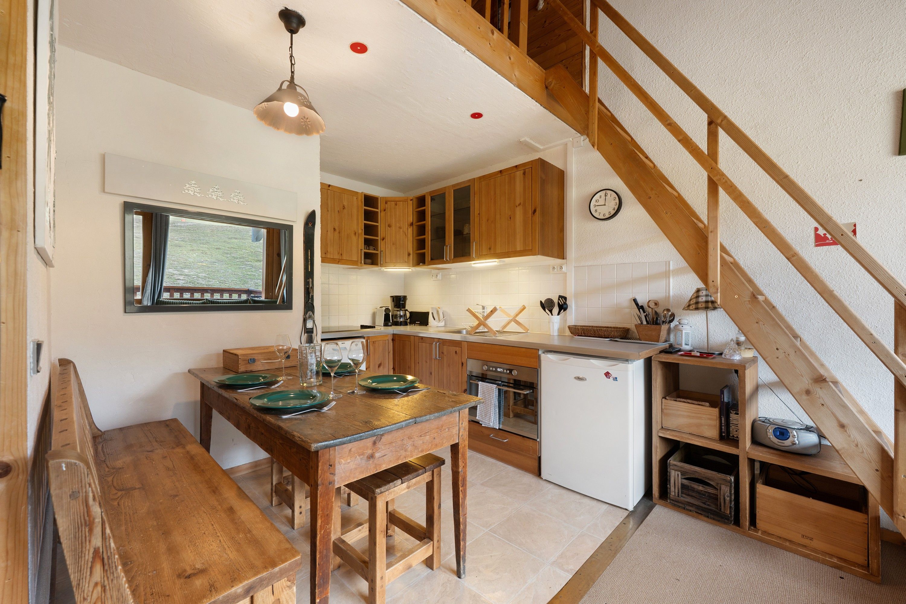 Une cuisine avec des meubles en bois, une table à manger avec des assiettes vertes, une horloge au mur et un escalier en bois.