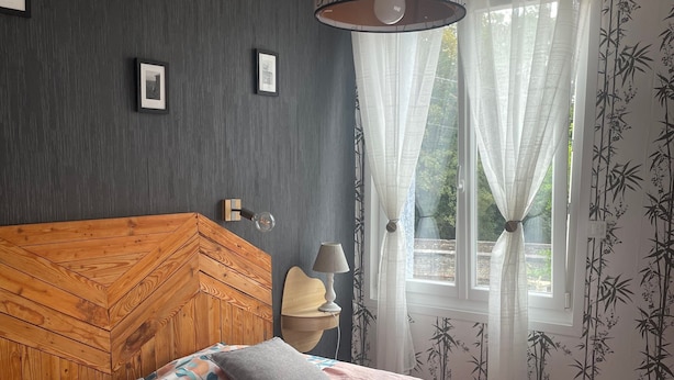 Een slaapkamer met een houten hoofdbord, een bed met een roze sprei, twee kussens, een bedlampje en een raam met dunne gordijnen.