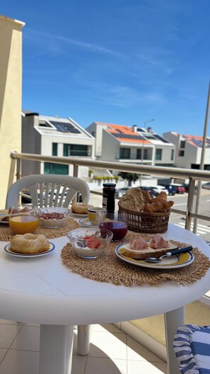 A table set with breakfast items including bread, pastries, and a drink, on a balcony with a view of buildings.
