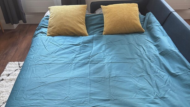 A bed with a blue bedspread and two yellow pillows, a white rug, and a window with curtains.