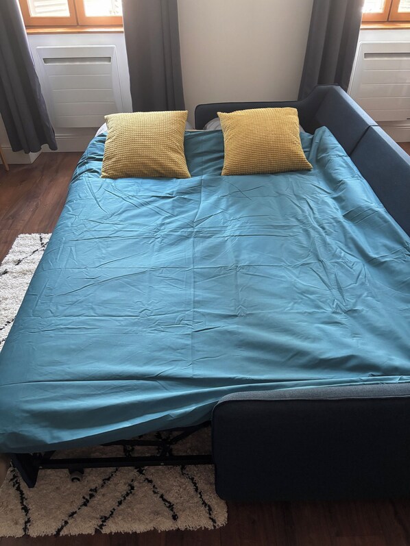 A bed with a blue bedspread and two yellow pillows, a white rug, and a window with curtains.