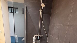 A modern bathroom with a glass shower enclosure, dark tiled walls, and a visible bottle of shampoo on the floor.