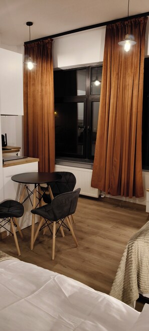 A compact room with a dining table and chairs, a window with curtains, a radiator, and a bed with a white cover.