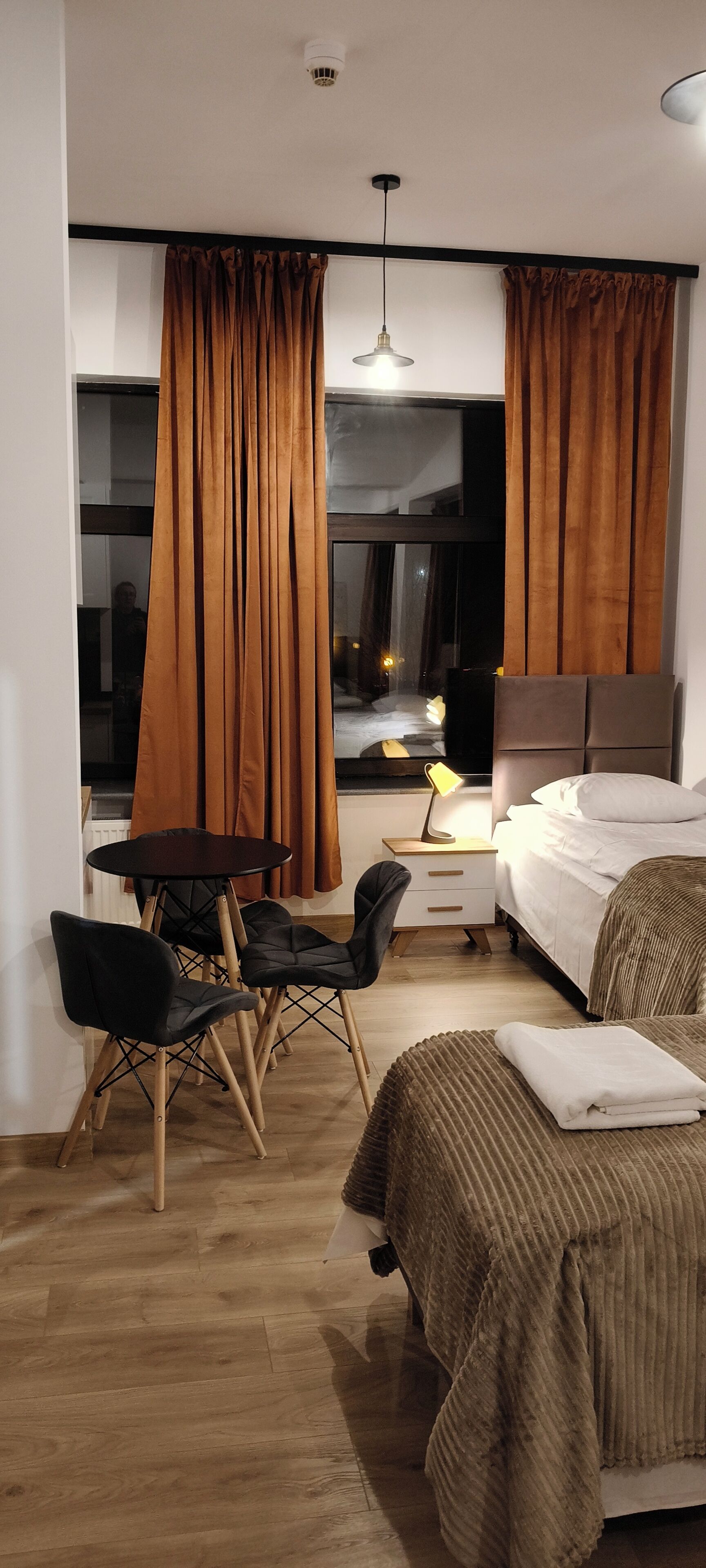 Una habitación de hotel moderna con cama, escritorio con sillas, mesita de noche con lámpara y ventanas amplias con cortinas.