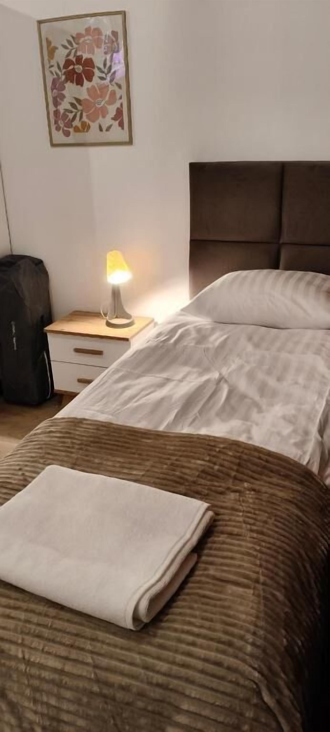 A neatly made bed with a folded towel, a bedside lamp, and a floral artwork on the wall.