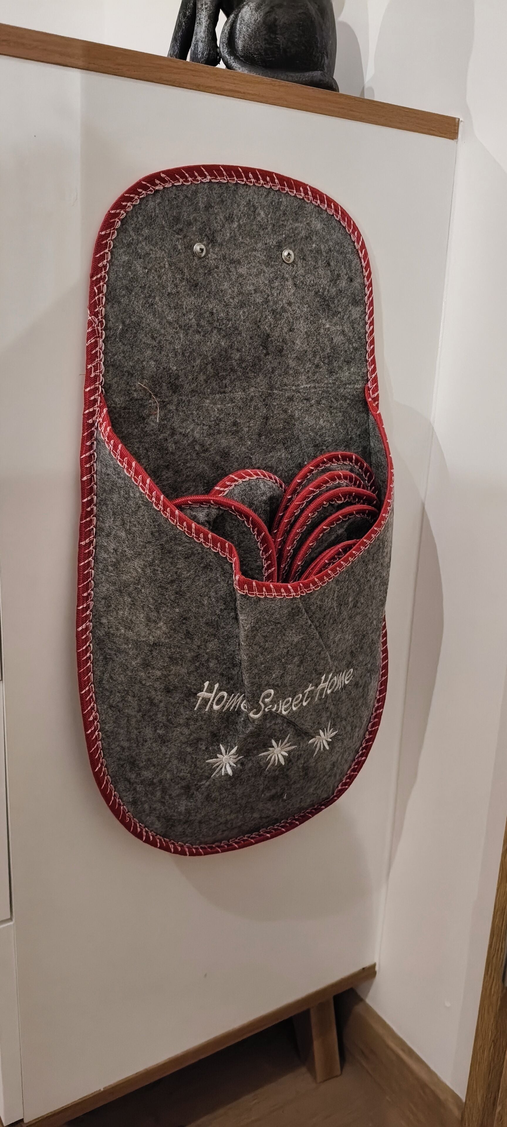 Een grijze opbergmand, bekleed met rood accentleer, met de tekst 'Home Sweet Home' en decoratieve sterren, bevestigd aan een witte kast.