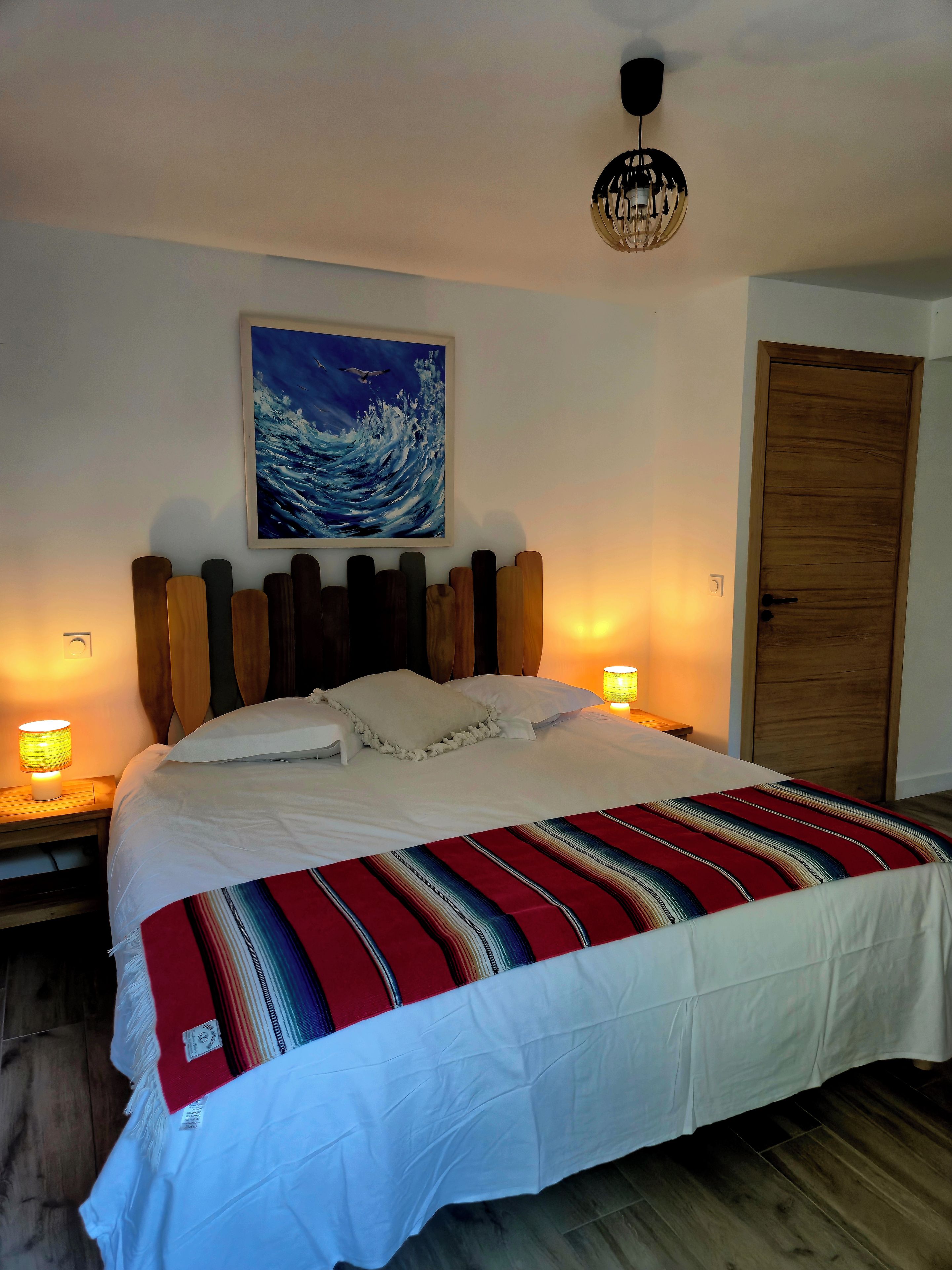 Een bed met een gestreepte deken, twee bedlampjes, een schilderij van golven en een houten deur.