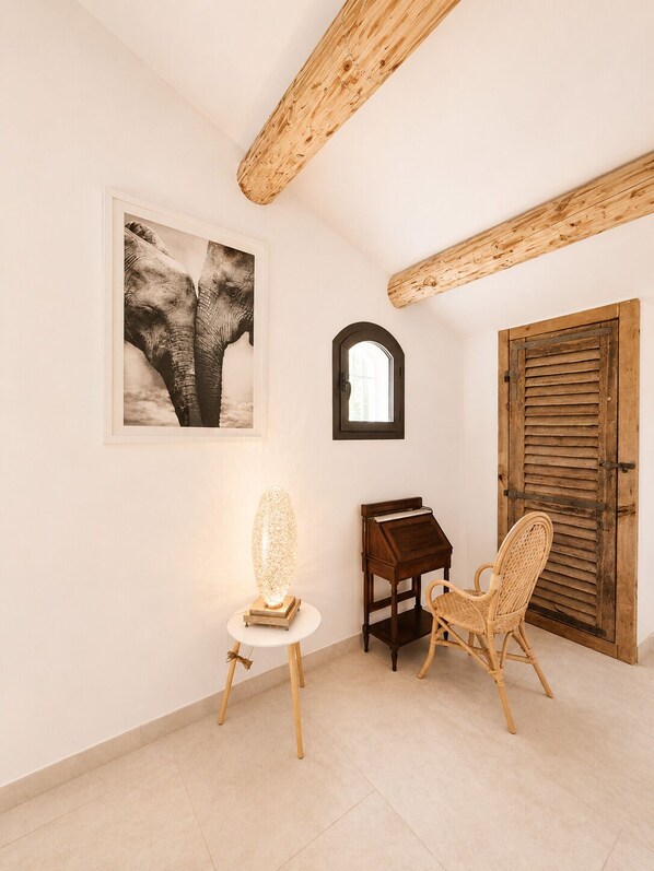 Une pièce avec une poutre en bois au plafond, un tableau représentant un éléphant encadré, une lampe de table, un piano et une chaise en osier.