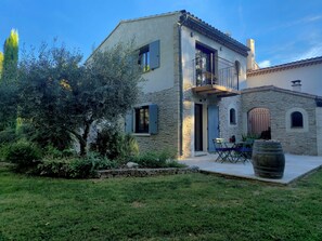 A two-story stone building with a balcony, a stone wall, and a grassy yard with a barrel.