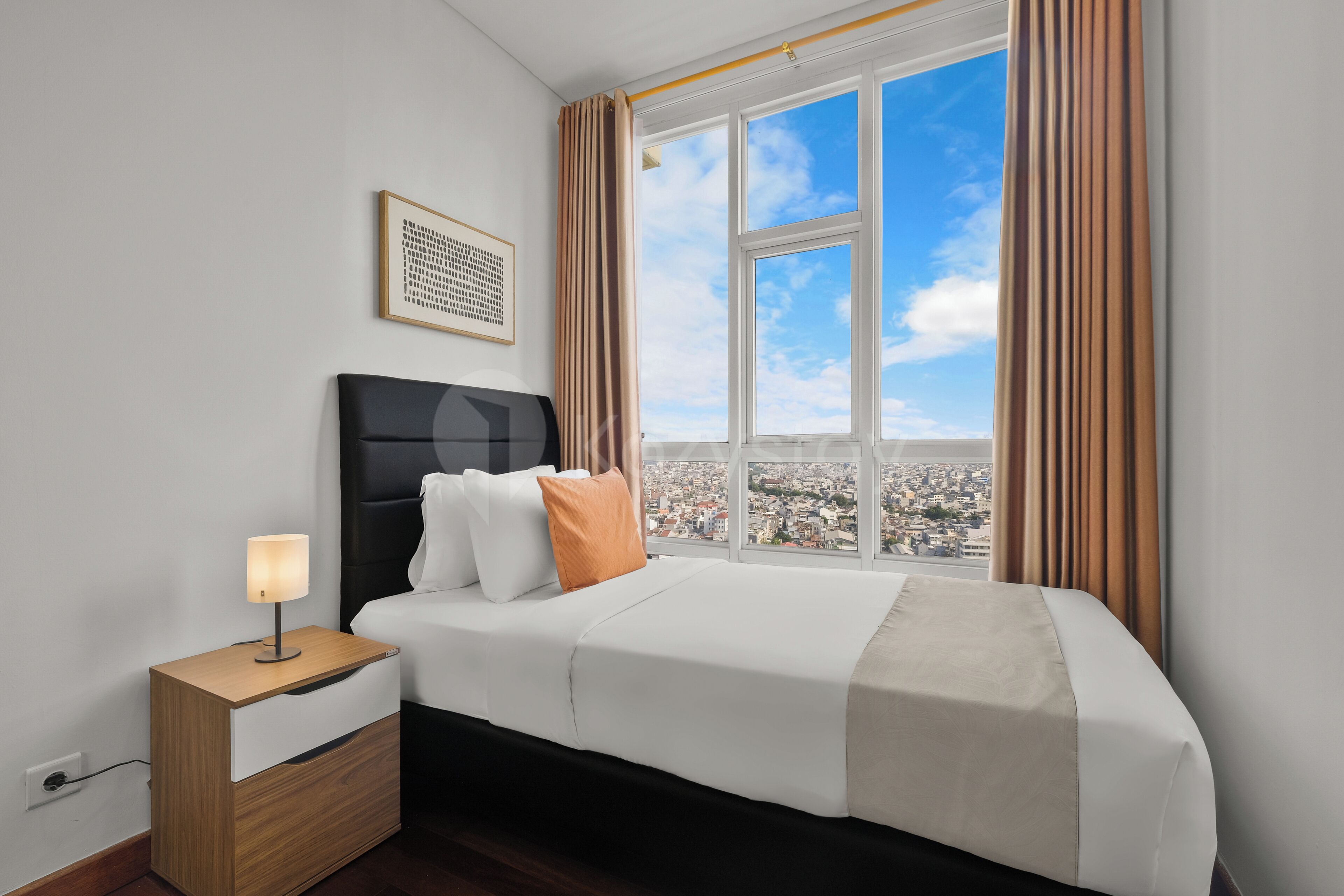 Habitación de hotel con cama, mesita de noche, lámpara, cuadro enmarco y ventana con cortinas.