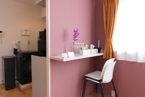 A room with a desk, a chair, a washing machine, and a small shelf with items.