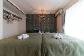 A room with two beds, a patterned wall, a shelf with storage boxes, and a hanging light fixture.